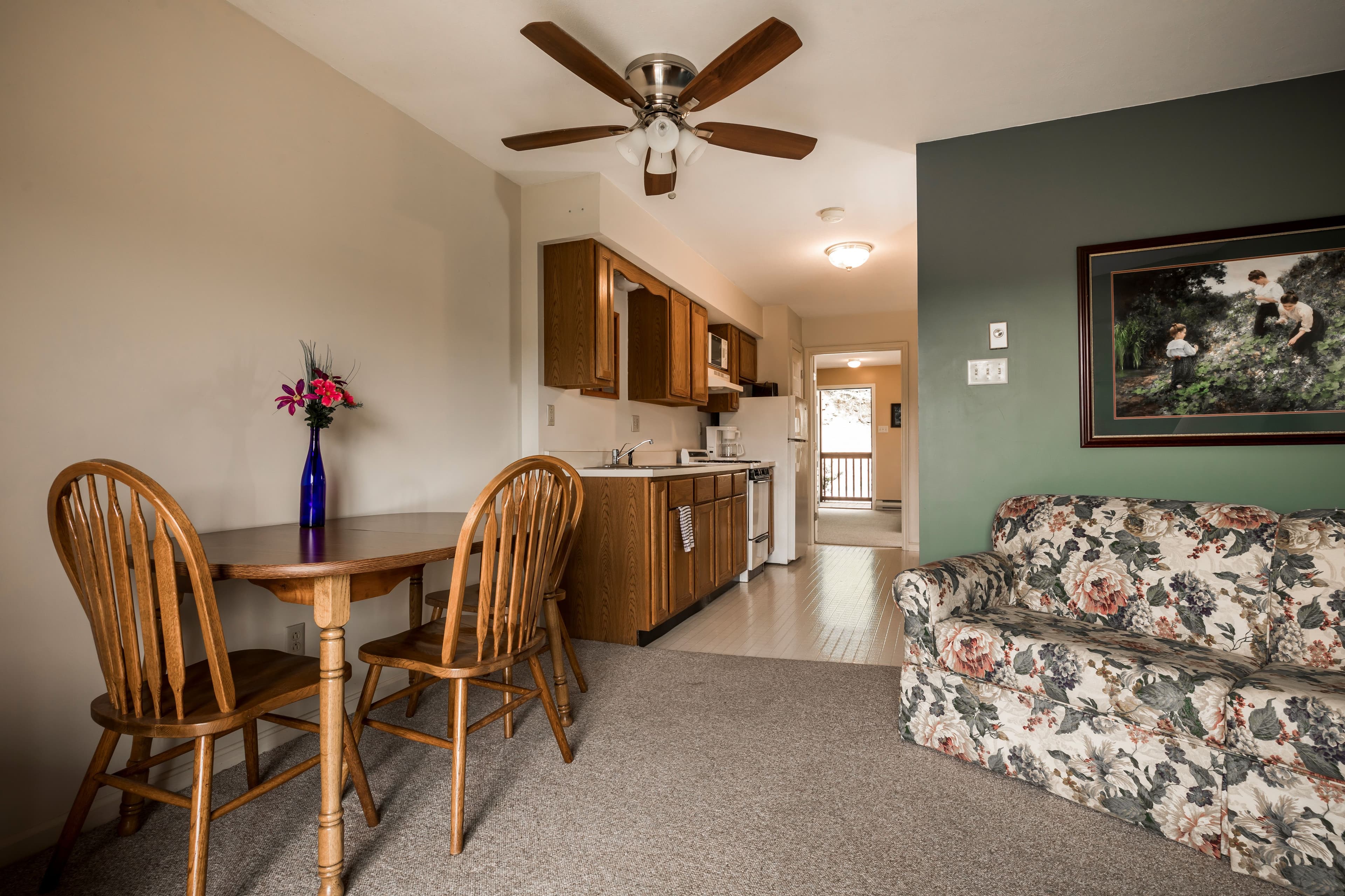 A cozy dining area with a wooden table and chairs, a floral couch, and a kitchenette in the background.