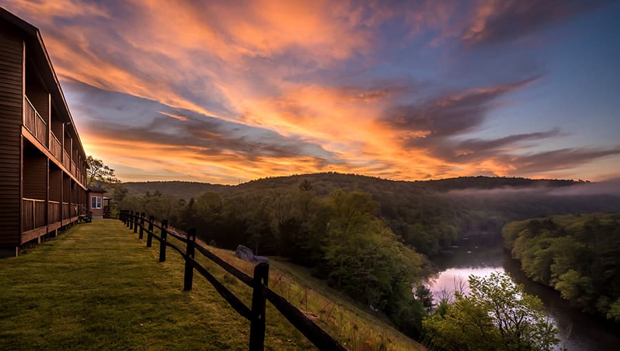 A tranquil riverside view at sunset, with a wooden building and lush greenery in the foreground.