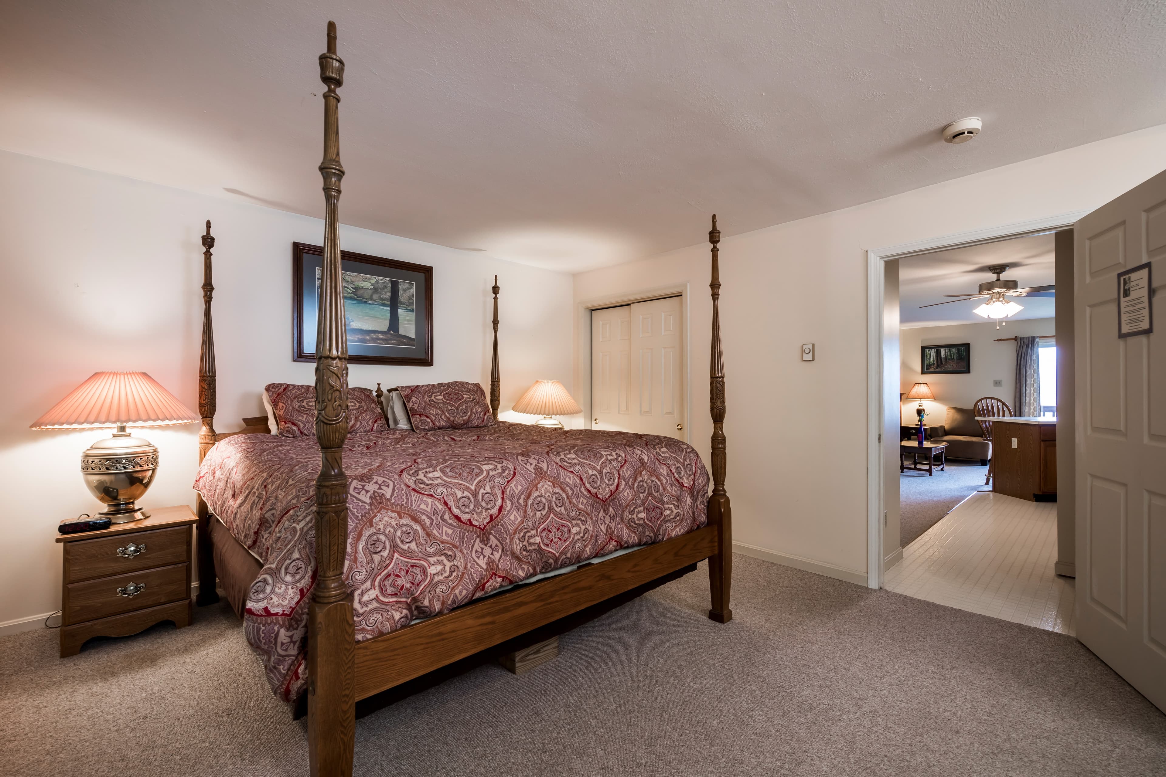 A cozy bedroom features a four-poster bed, bedside lamp, and an open door leading to another room.