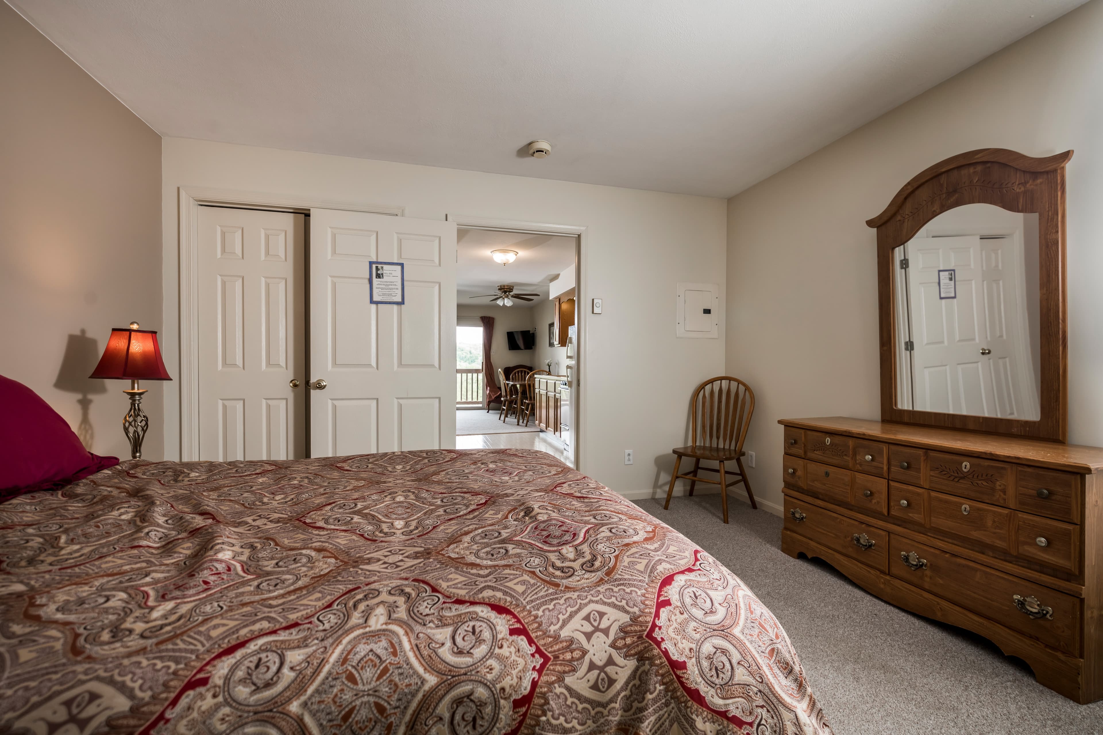 View of bedroom in typical family suite from wall to parking lot, with queen-sized bed, closet and full-sized chest of drawers, looking towards door to kitchen and through kitchen into the living area