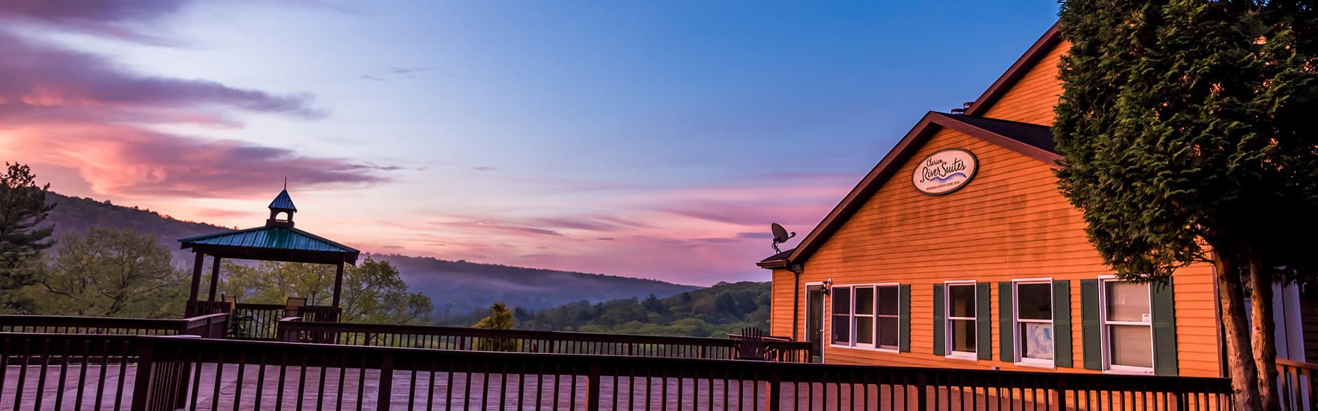 A rustic lodge with a wooden deck overlooks a vibrant sunrise and misty hills.