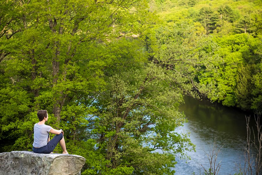 A person sits on a rock overlooking a serene river surrounded by lush green trees.