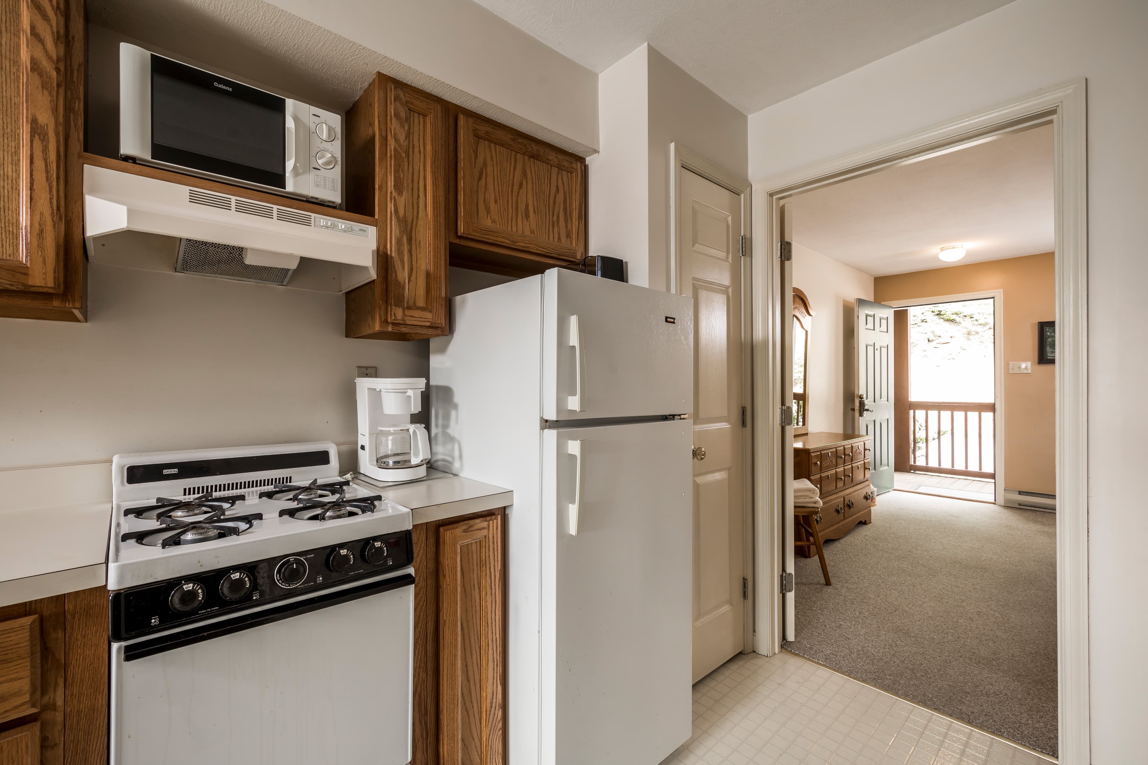 A small kitchen featuring a stove, refrigerator, and microwave, adjacent to an entryway leading to a living area.