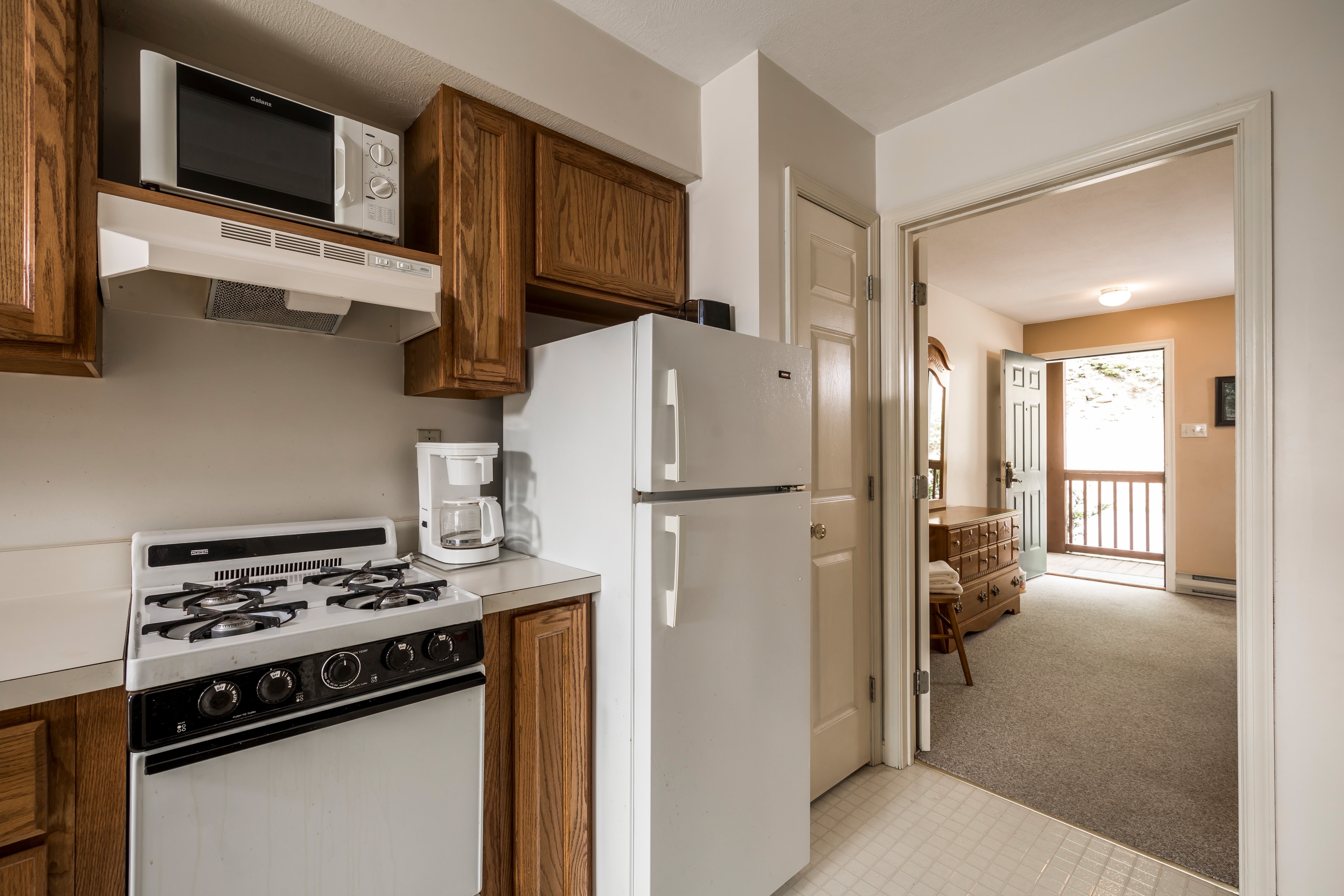 A small kitchen featuring a stove, refrigerator, and microwave, adjacent to an entryway leading to a living area.