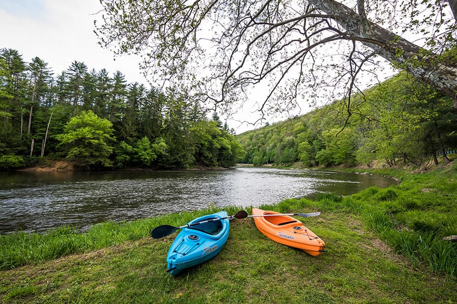 Two kayaks, one blue and one orange, are resting on the grassy bank beside a serene river surrounded by lush greenery.