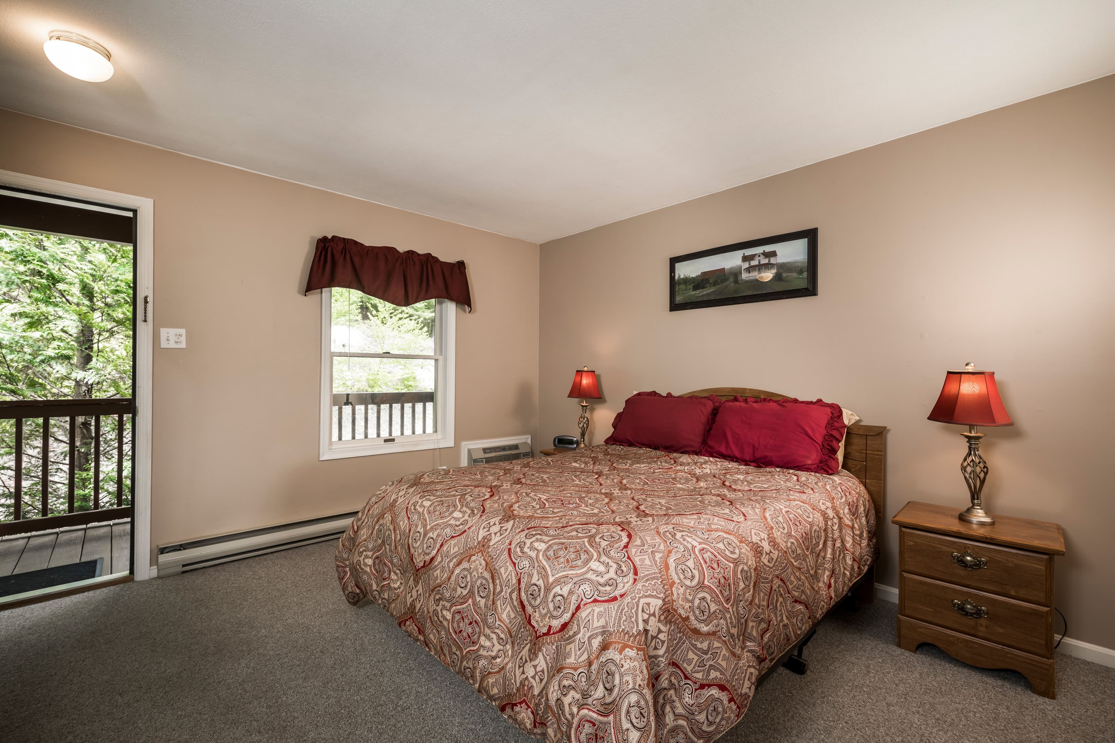 View of bedroom in typical family suite from door to kitchen, with queen-sized bed with two sets of pillows and quilt, two bed tables with lamps, overhead fan and light, wall mounted air-conditioner and main entrance to parking lot