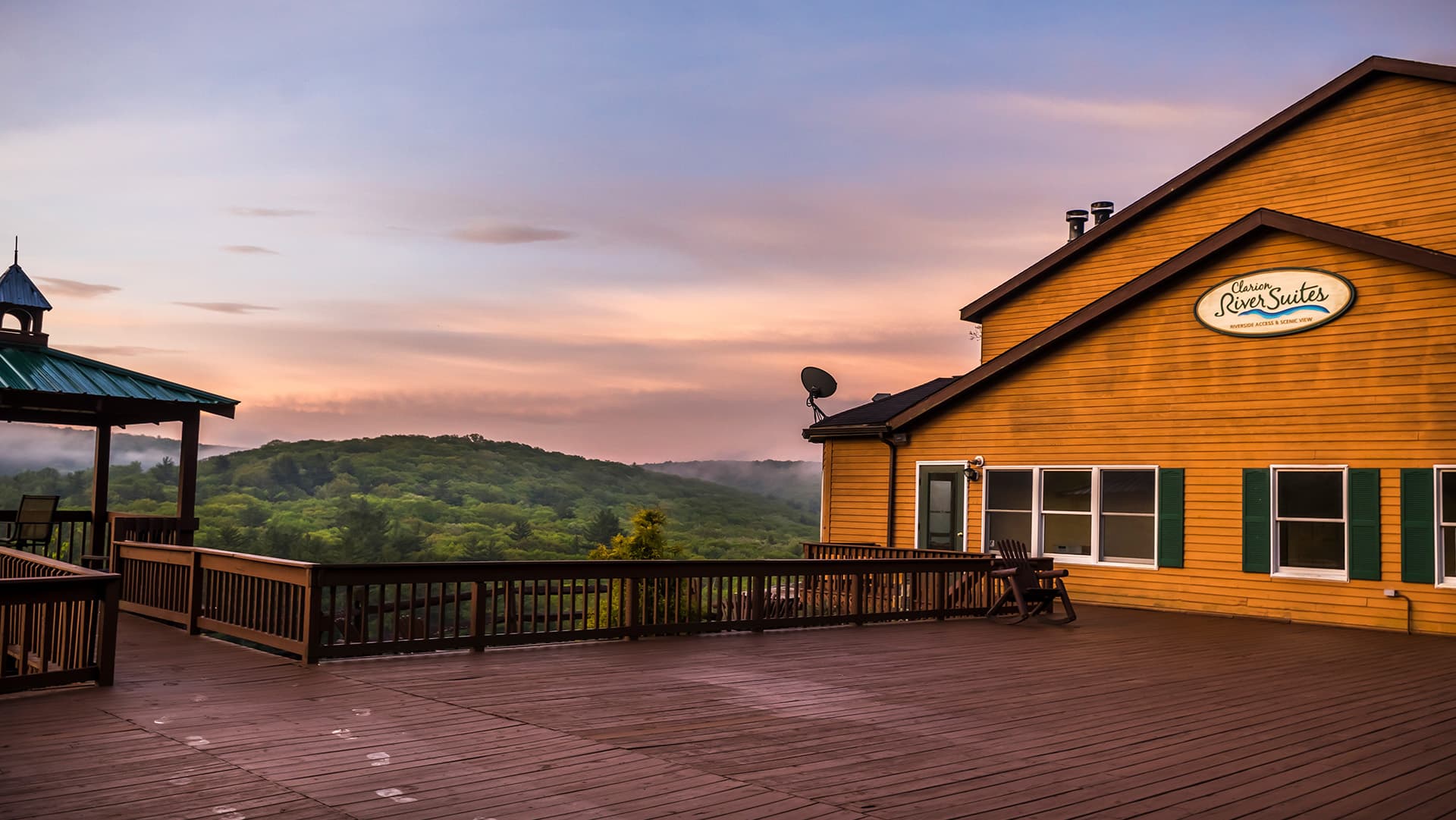 A yellow lodge with a green roof overlooks a misty, wooded landscape at sunset.