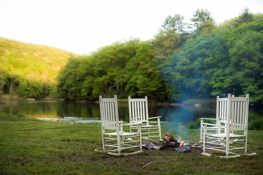 Four white rocking chairs are arranged around a small campfire by a serene lake surrounded by lush greenery.