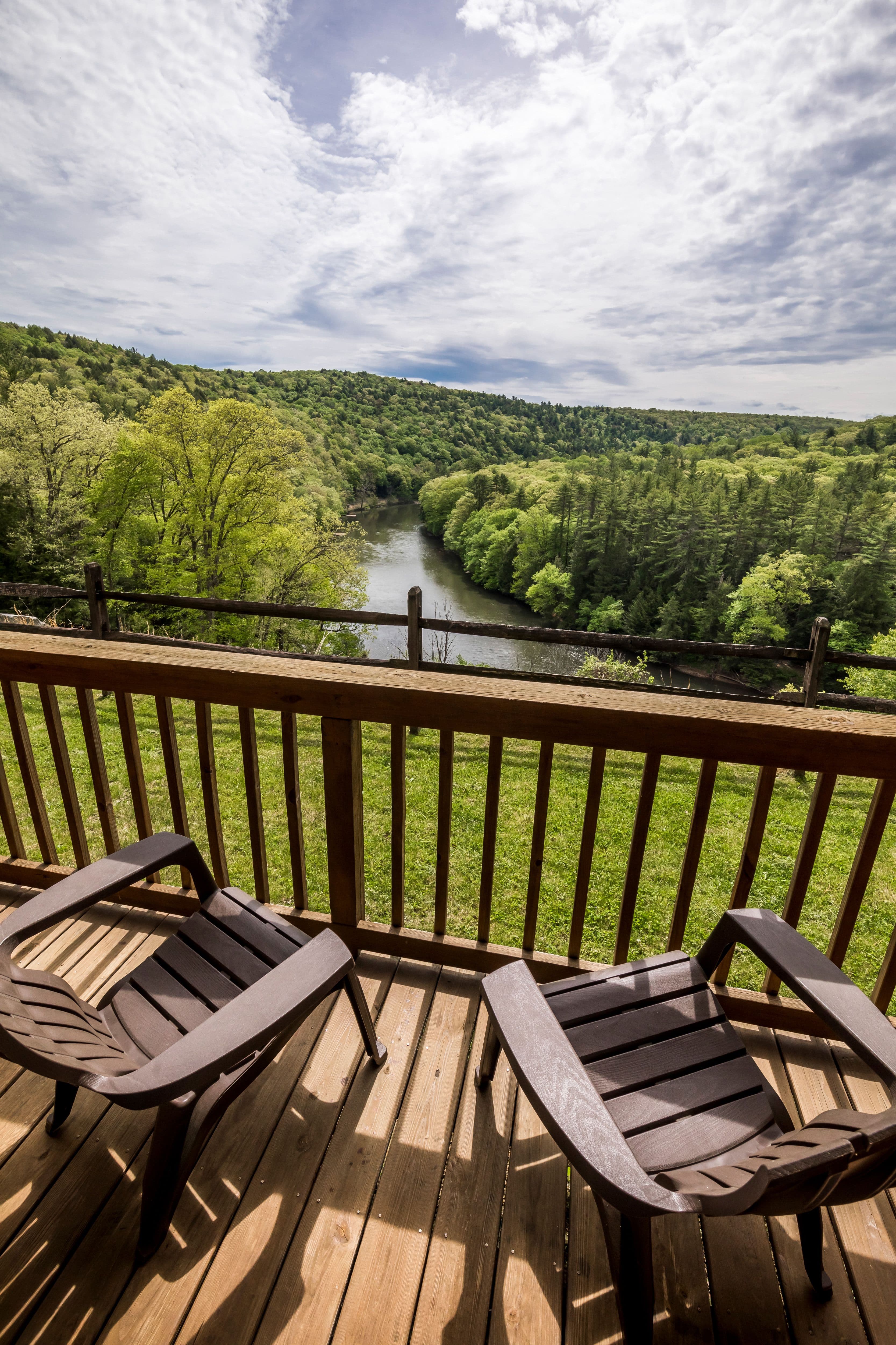 Two wooden chairs overlook a serene river surrounded by green trees under a partly cloudy sky.