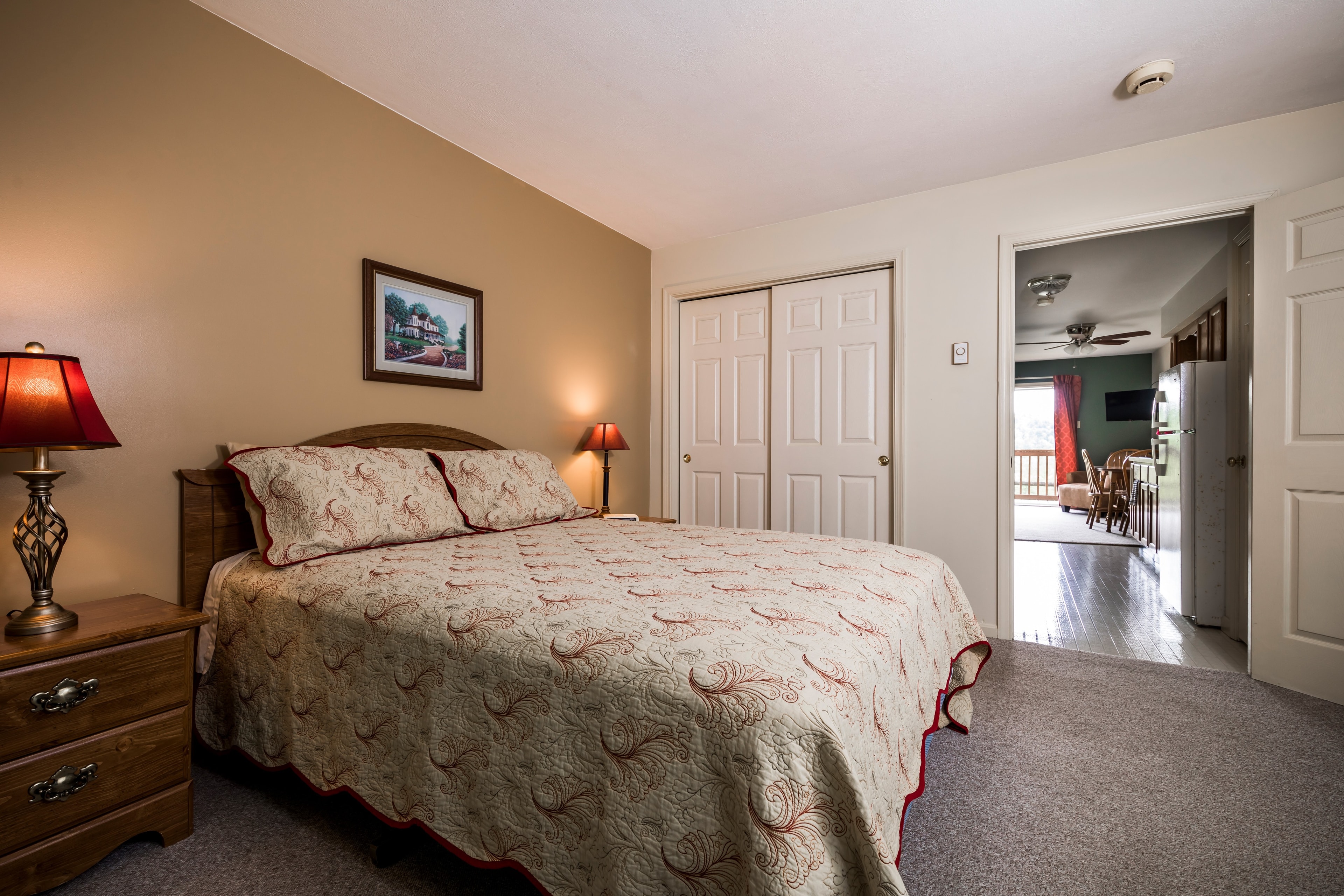 A cozy bedroom featuring a bed with a patterned quilt, bedside lamp, and a doorway leading to another room.