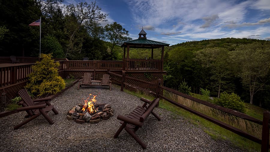 A fire pit surrounded by wooden chairs, with a gazebo and trees in the background under a partly cloudy sky.