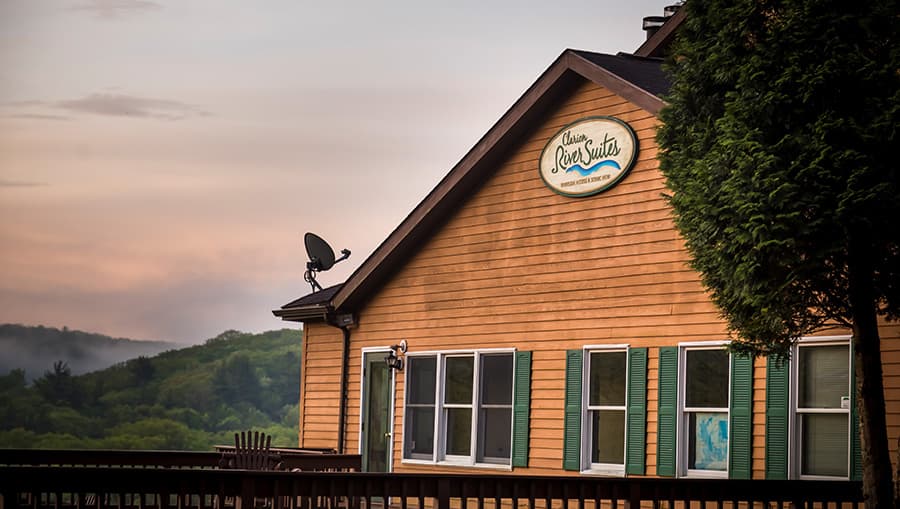 A wooden building with green shutters and a sign reading "Clare's River Suites" beneath a hazy sky.